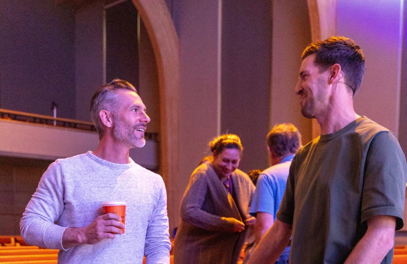 Two men talking and laughing in the auditorium, one holding a red cup