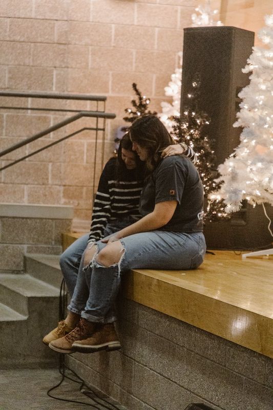 Two friends hugging on the edge of a stage beside white Christmas trees strung with lights