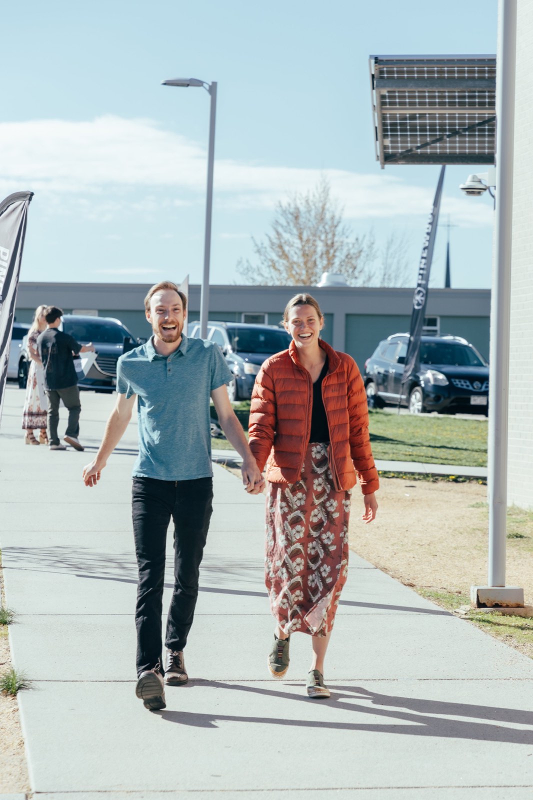 A couple holding hands and laughing as they walk up the sidewalk toward the Sunday gathering, Pinewood flags and parked cars behind them