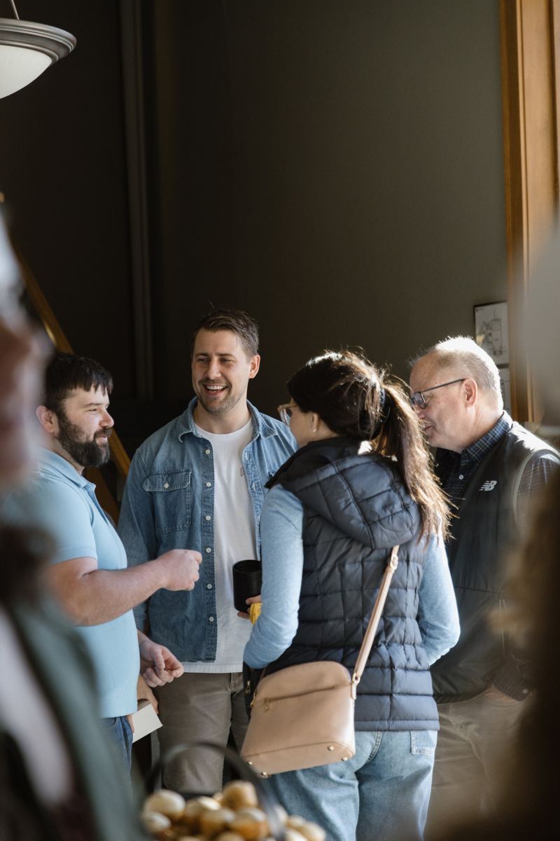 Adults of different ages talking and smiling by a window, a grey-haired man in glasses among them
