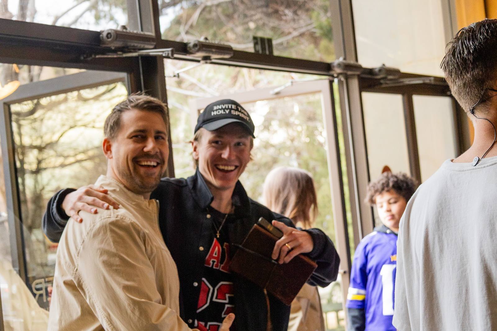 Two friends laughing with an arm around each other by the sunlit lobby doors, one in a cap holding a leather-bound book