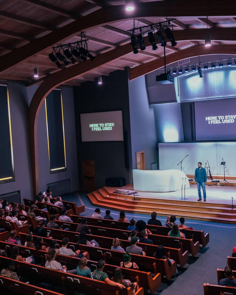 A full sanctuary seen from the balcony: rows of people in wooden pews facing a speaker on a lit stage, with screens reading Here to Stay: I Feel Used