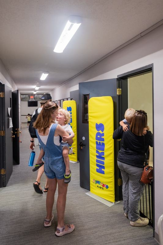 A parent carrying her toddler past the Hikers classroom banner at check-in while another parent hugs her child at the door