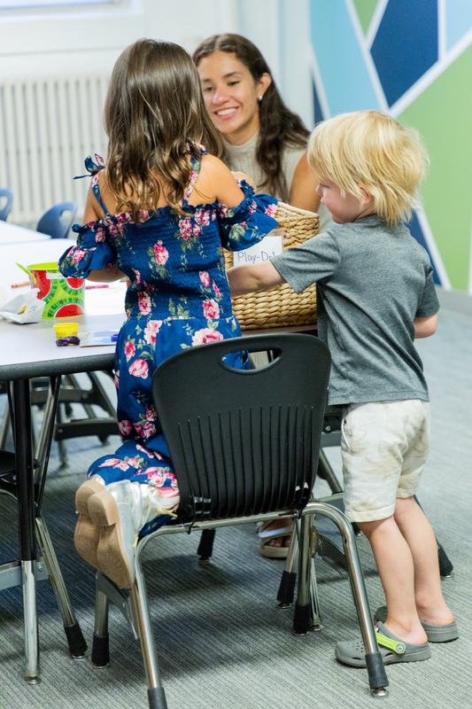 A smiling leader at a classroom table with a girl in a floral dress and a blond boy, play dough on the table