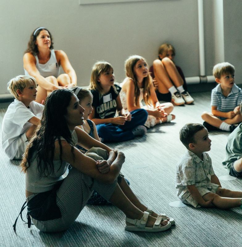 Two leaders sitting on the floor with a group of kids, everyone looking toward the front of the room