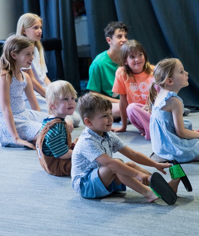 Kids sitting on the floor in front of a dark curtain, a smiling boy in the front row