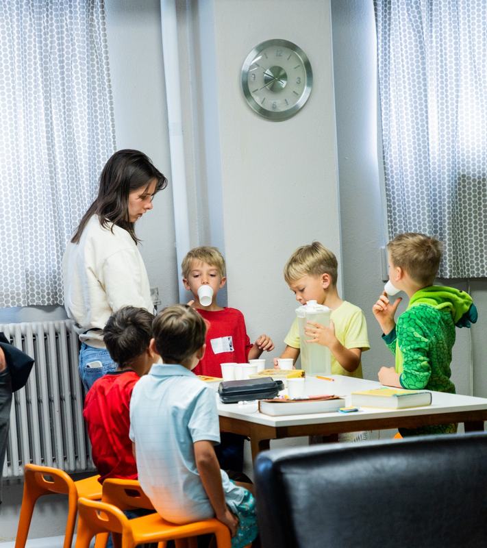 Boys drinking from paper cups around a classroom table while a leader stands beside them