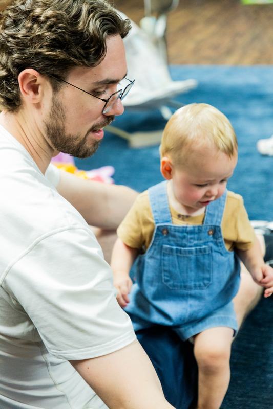 A man in glasses sitting on the rug beside a baby in denim overalls