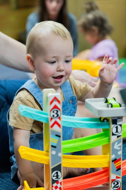A smiling baby waving a hand over a colorful toy car ramp