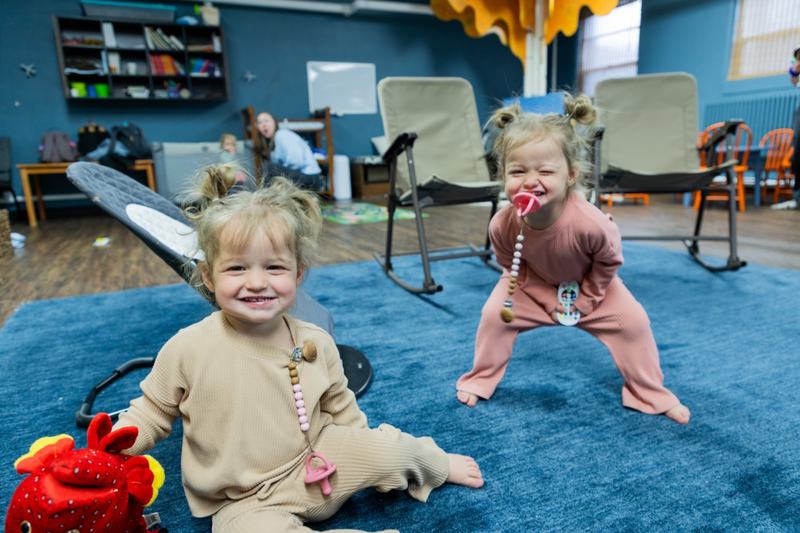 Two toddlers on a blue rug, one grinning at the camera and one making a playful face
