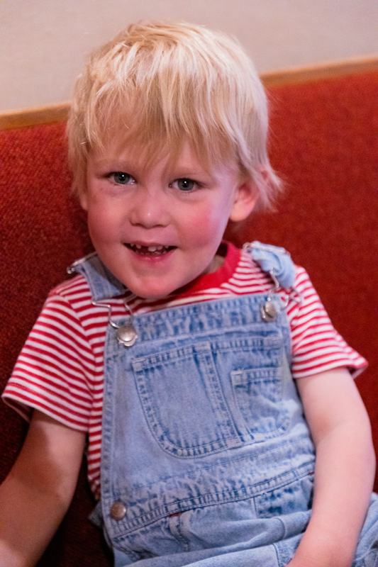 A smiling blond boy in a red striped shirt and denim overalls