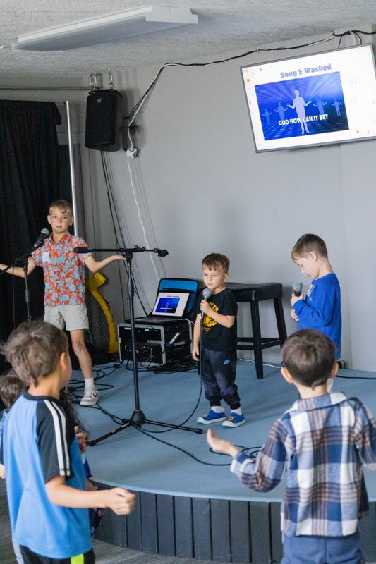 Boys singing into microphones on a small stage, with the song on a screen behind them
