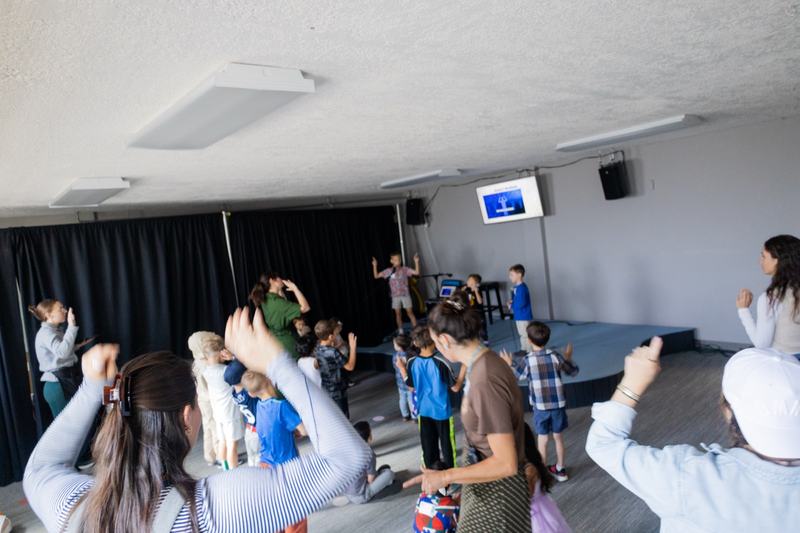 Kids and leaders with their hands raised during worship in the PNWD Kids room, a child leading from the small stage