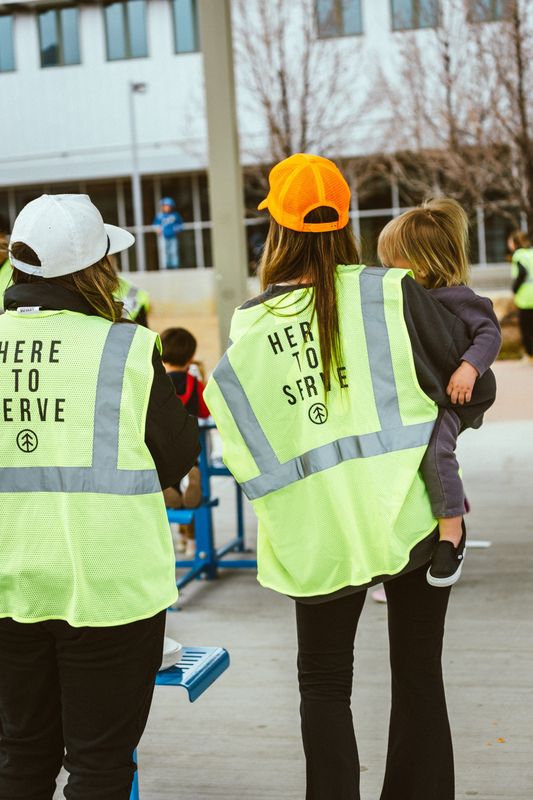 Two volunteers seen from behind in yellow safety vests printed Here to serve, one carrying a small child