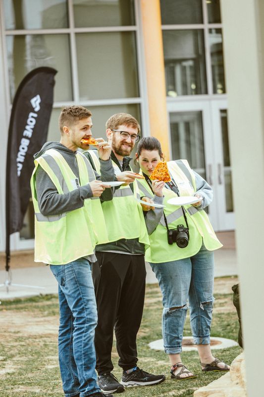 Three volunteers in yellow safety vests eat pizza beside a Love Boulder flag