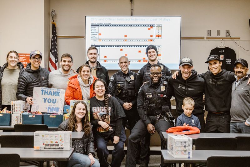 Volunteers and uniformed police officers pose together in a briefing room beside boxes of gifts and a hand-lettered thank-you sign
