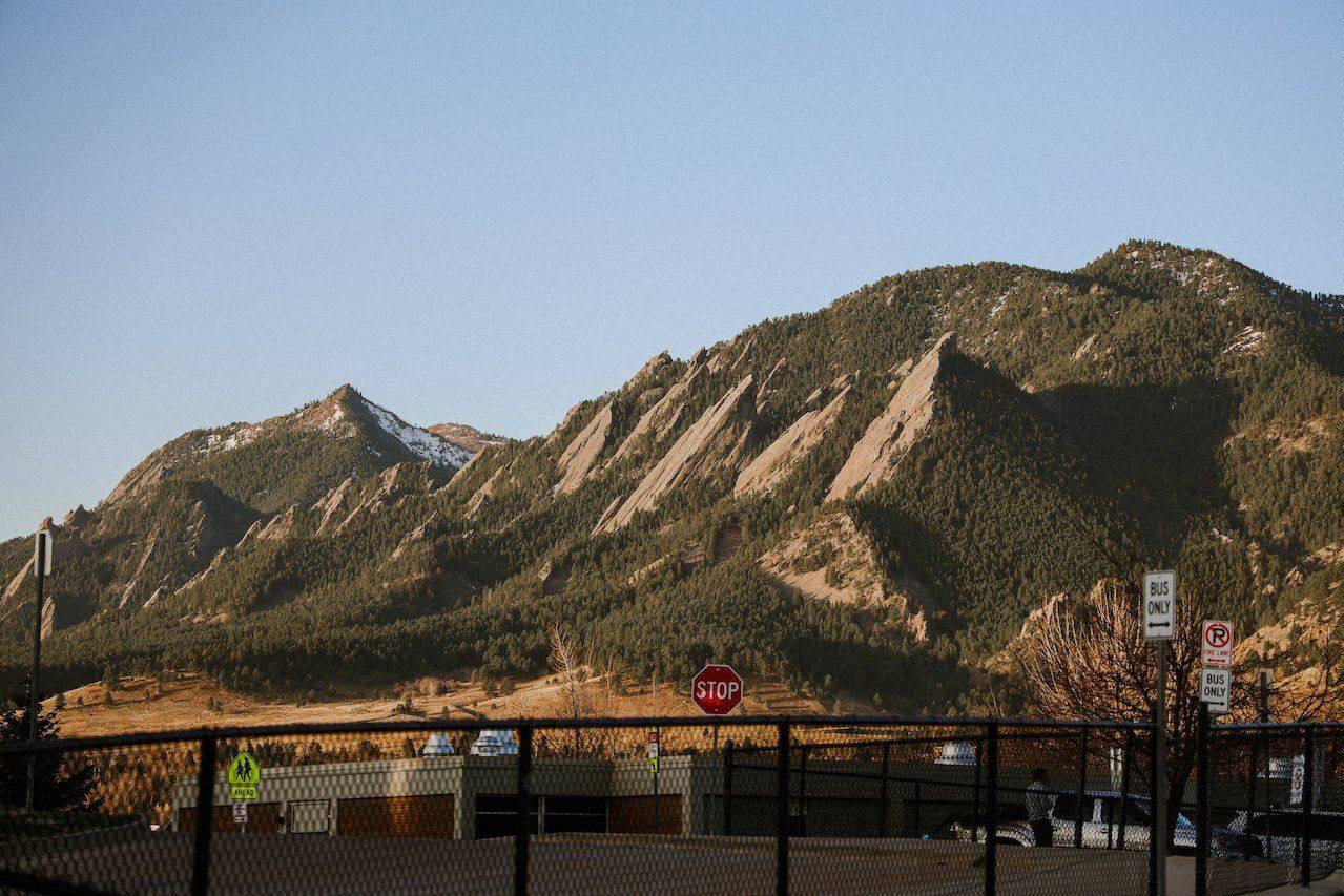 The Flatirons above Boulder in late afternoon light, seen across a fenced lot with a stop sign