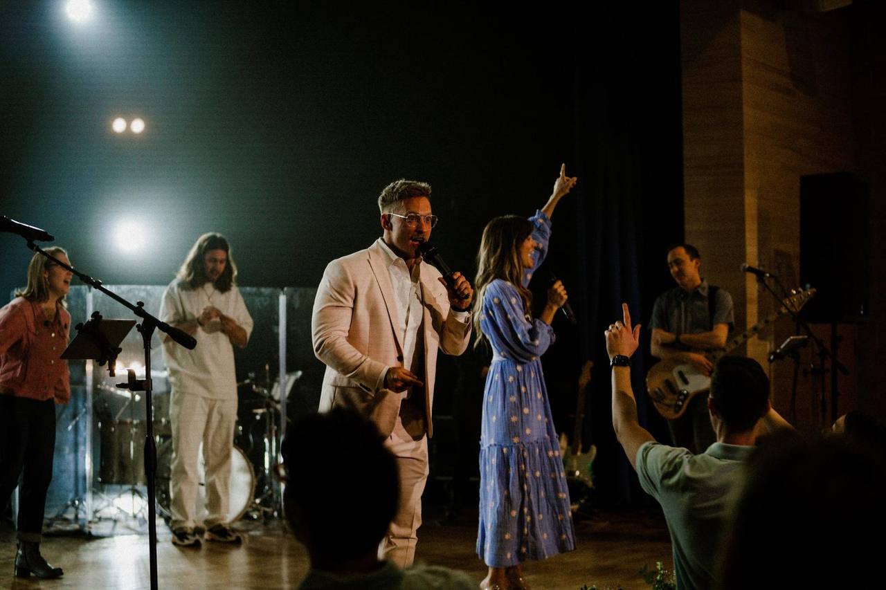 The worship team leads from a lit stage on a Sunday, one singer with a hand raised