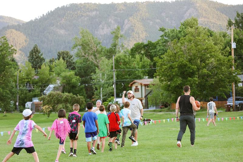 Kids and two adults crossing a grassy park toward a game, one adult carrying a basketball, wooded foothills behind them
