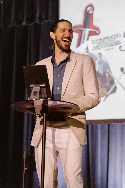 A laughing man in a light linen suit speaking on stage from a small round lectern with a tablet on it