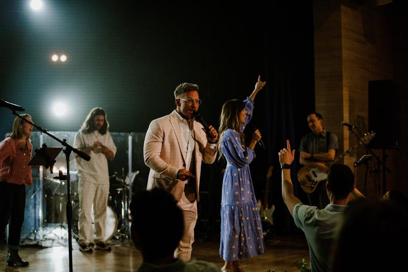 A man in a cream suit and a woman in a blue dress singing on stage with the band, a hand raised in the room