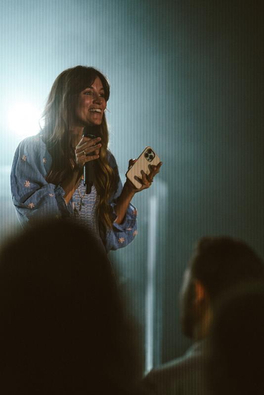 A woman in a blue embroidered top smiling as she speaks into a microphone, a phone in her other hand, lit from behind