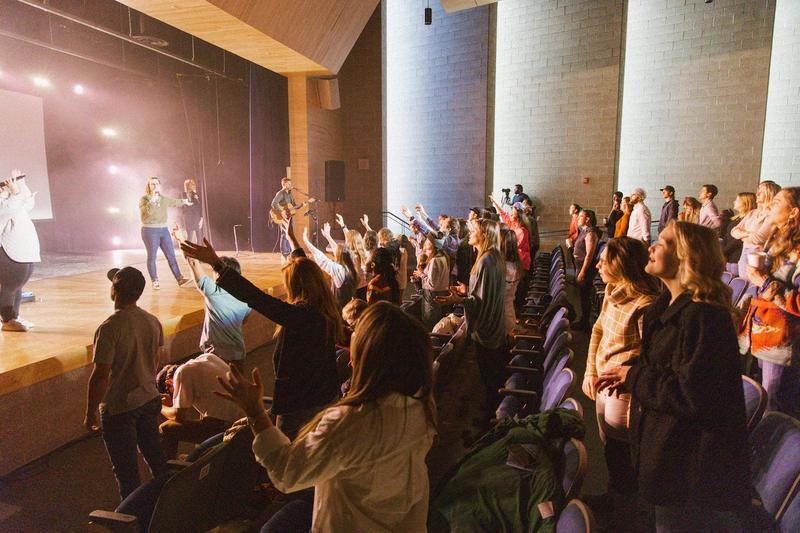 An auditorium on its feet in worship, hands raised toward a band on a stage lit pink and white