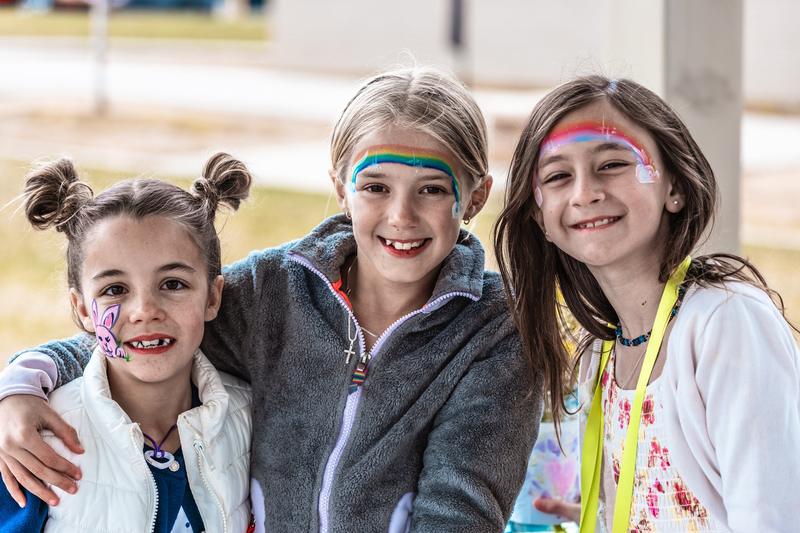Three smiling girls with rainbow face paint, arms around each other