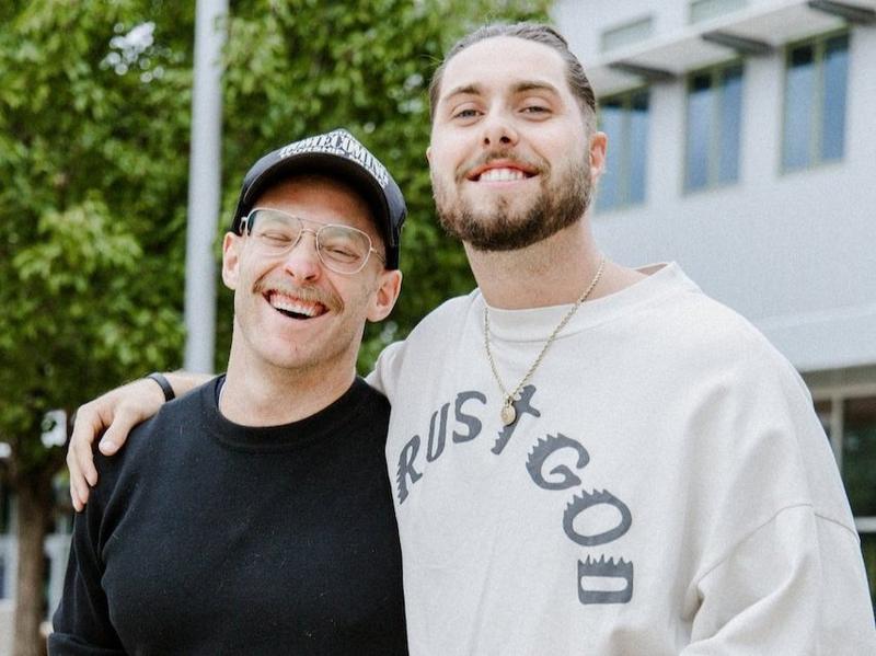Two men grinning arm in arm outdoors, trees and a white building behind them
