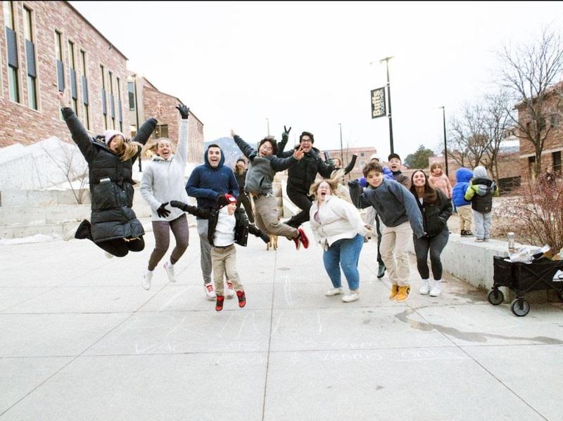 Adults and kids jumping in the air for a photo on a snowy plaza between brick buildings