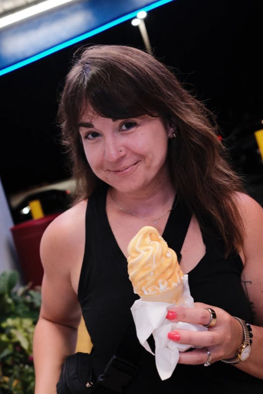 Kayla Slaughter, brown hair with bangs, smiling and holding a butterscotch-dipped ice cream cone at night