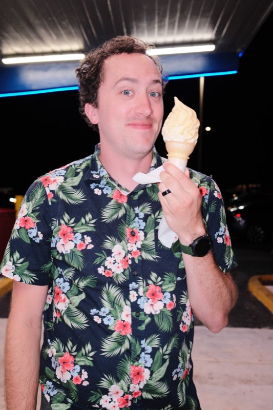 Keegan Velasquez in a floral shirt, holding up a vanilla ice cream cone under a lit awning at night