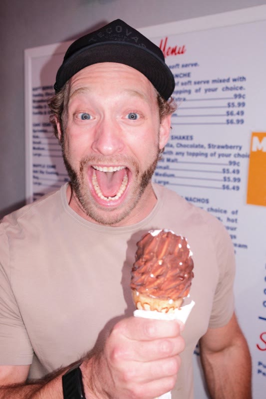 Matthew Slaughter in a black cap, laughing with his mouth wide open and holding up a chocolate-dipped ice cream cone