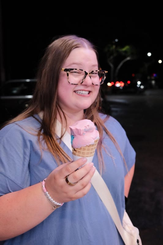 Melody Sebald in glasses and a blue top, smiling and holding a pink ice cream cone at night